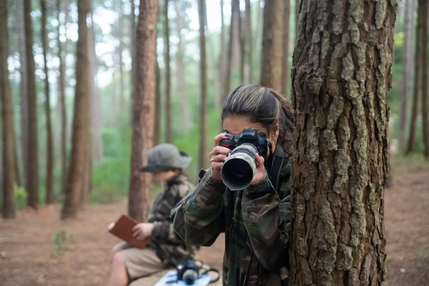 Eine konzentrierte Mutter im Mantel und mit Pferdeschwanz fotografiert mit einer Kamera im Wald; ihr unscharfer Sohn ist im Hintergrund zu sehen. Thema: Natur und Freizeit.